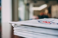 A stack of folded white napkins with a red letter 'B' printed on them, placed on a dark wooden surface. The background appears blurred, suggesting an indoor setting with soft lighting.