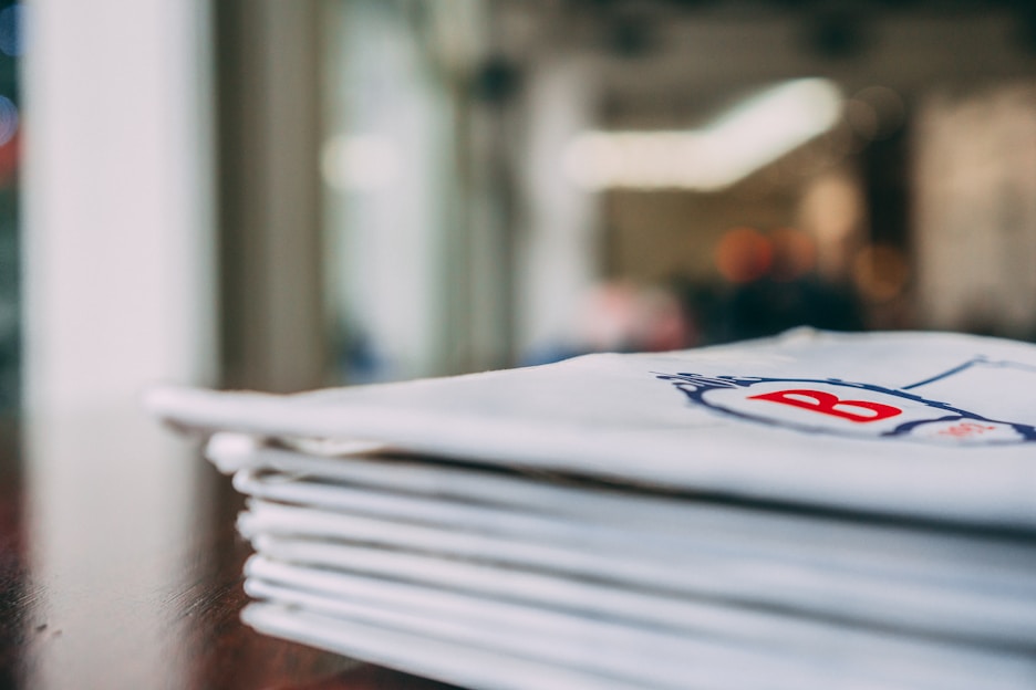 A stack of folded white napkins with a red letter 'B' printed on them, placed on a dark wooden surface. The background appears blurred, suggesting an indoor setting with soft lighting.
