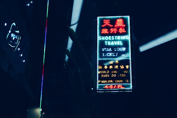 A neon sign with both English and Chinese characters illuminates against a dark background. The sign advertises a travel agency with services such as visa, tour, and ticket assistance. The surrounding area is dimly lit, with faint reflections visible.
