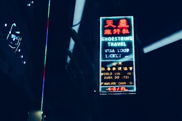 A neon sign with both English and Chinese characters illuminates against a dark background. The sign advertises a travel agency with services such as visa, tour, and ticket assistance. The surrounding area is dimly lit, with faint reflections visible.