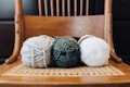 Three balls of yarn are placed side by side on a woven chair. The yarn colors are gray, dark green, and white, with the textured chair adding a rustic and cozy appeal. The background consists of a wooden chair back and darker tones, creating a warm atmosphere.