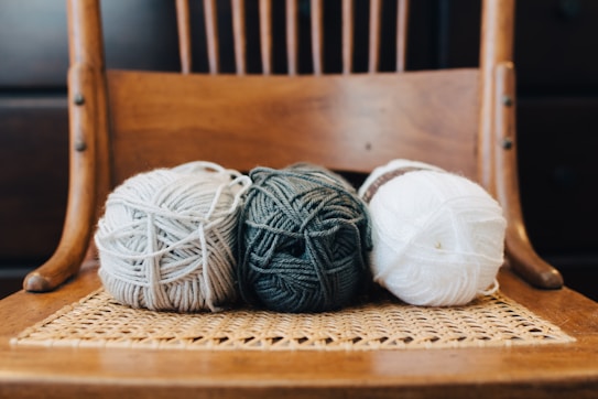 Three balls of yarn are placed side by side on a woven chair. The yarn colors are gray, dark green, and white, with the textured chair adding a rustic and cozy appeal. The background consists of a wooden chair back and darker tones, creating a warm atmosphere.