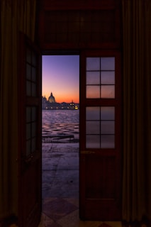 An elegant doorway opens to a picturesque view of a calm body of water during sunset. In the distance, the silhouette of an iconic domed building is visible against a vibrant sky transitioning from shades of orange to deep blue. The scene is framed by the rich wood of the door and muted curtains inside.