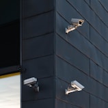 Three security cameras are mounted on the exterior of a modern building with dark paneling. The cameras are positioned at different angles to cover a wide area. The building corner is sharply defined, with a section of glass window partially visible.