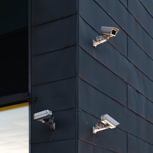 Three security cameras are mounted on the exterior of a modern building with dark paneling. The cameras are positioned at different angles to cover a wide area. The building corner is sharply defined, with a section of glass window partially visible.