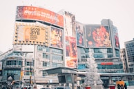 An urban cityscape featuring a bustling commercial area with large advertisements and billboards. Prominent brand logos like Coca-Cola are displayed on the large screens. The area is lively with various shops and brands like Cineplex, Adidas, and Winners visible. There is a decorative sculpture resembling a Christmas tree made of white spherical structures in the foreground.