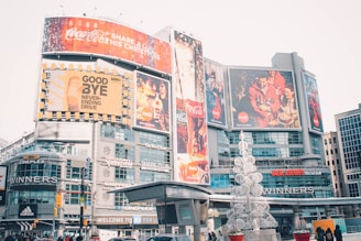 An urban cityscape featuring a bustling commercial area with large advertisements and billboards. Prominent brand logos like Coca-Cola are displayed on the large screens. The area is lively with various shops and brands like Cineplex, Adidas, and Winners visible. There is a decorative sculpture resembling a Christmas tree made of white spherical structures in the foreground.
