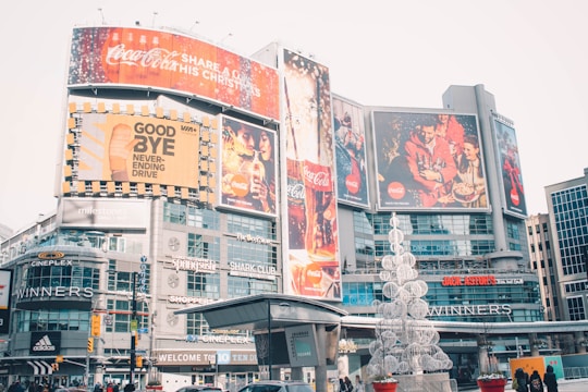 An urban cityscape featuring a bustling commercial area with large advertisements and billboards. Prominent brand logos like Coca-Cola are displayed on the large screens. The area is lively with various shops and brands like Cineplex, Adidas, and Winners visible. There is a decorative sculpture resembling a Christmas tree made of white spherical structures in the foreground.