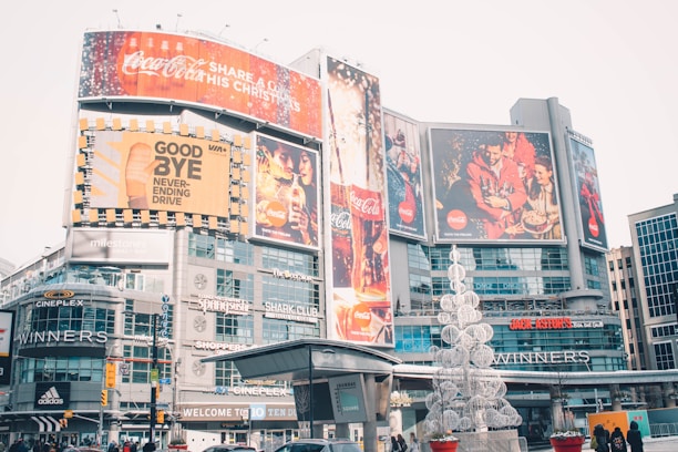 An urban cityscape featuring a bustling commercial area with large advertisements and billboards. Prominent brand logos like Coca-Cola are displayed on the large screens. The area is lively with various shops and brands like Cineplex, Adidas, and Winners visible. There is a decorative sculpture resembling a Christmas tree made of white spherical structures in the foreground.