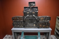 A collection of vintage military communication equipment is stacked neatly on a gray table. The devices are covered in dials, knobs, and connectors, indicating their complex functionalities. The room has wooden paneling and a tiled floor with a repetitive black and white pattern. A green chair is positioned in front of the table.