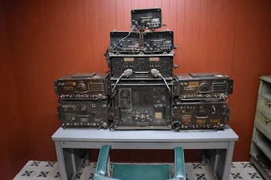A collection of vintage military communication equipment is stacked neatly on a gray table. The devices are covered in dials, knobs, and connectors, indicating their complex functionalities. The room has wooden paneling and a tiled floor with a repetitive black and white pattern. A green chair is positioned in front of the table.