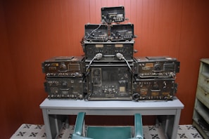 A collection of vintage military communication equipment is stacked neatly on a gray table. The devices are covered in dials, knobs, and connectors, indicating their complex functionalities. The room has wooden paneling and a tiled floor with a repetitive black and white pattern. A green chair is positioned in front of the table.