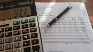 A close-up view of a financial document with printed figures and text, accompanied by a black pen resting on the paper. To the left, a digital calculator displays a number, suggesting it is used for calculations related to the document.