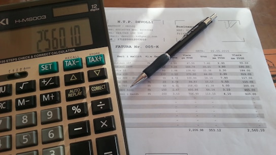 A close-up view of a financial document with printed figures and text, accompanied by a black pen resting on the paper. To the left, a digital calculator displays a number, suggesting it is used for calculations related to the document.