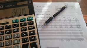 A close-up view of a financial document with printed figures and text, accompanied by a black pen resting on the paper. To the left, a digital calculator displays a number, suggesting it is used for calculations related to the document.