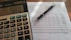 A close-up view of a financial document with printed figures and text, accompanied by a black pen resting on the paper. To the left, a digital calculator displays a number, suggesting it is used for calculations related to the document.