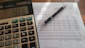 A close-up view of a financial document with printed figures and text, accompanied by a black pen resting on the paper. To the left, a digital calculator displays a number, suggesting it is used for calculations related to the document.