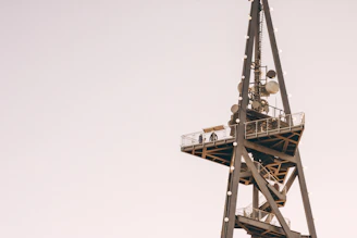 A tall communication tower with various satellite dishes and antennas attached. A viewing platform with a railing is present, and two silhouettes of people can be seen standing on it. The structure is made of metal and set against a clear, pale sky.