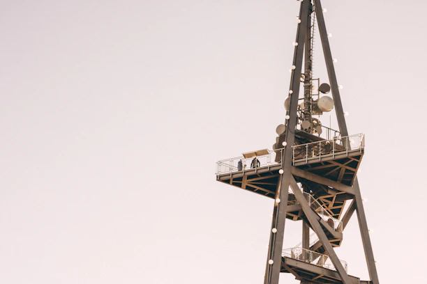 A tall communication tower with various satellite dishes and antennas attached. A viewing platform with a railing is present, and two silhouettes of people can be seen standing on it. The structure is made of metal and set against a clear, pale sky.