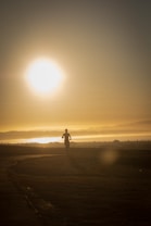 A person is jogging along a pathway during sunrise or sunset, with the sun low in the sky casting long shadows. The scene is tranquil, with a warm golden hue dominating the landscape. The horizon is visible with mist or low clouds adding a sense of depth.