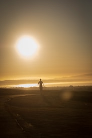 A person is jogging along a pathway during sunrise or sunset, with the sun low in the sky casting long shadows. The scene is tranquil, with a warm golden hue dominating the landscape. The horizon is visible with mist or low clouds adding a sense of depth.