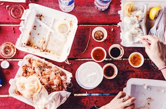 A red outdoor table is covered with various takeout food containers, some of which are empty or almost empty. There are disposable cups with sauces, lemon wedges, rice, and shrimp shells visible. Hands are using plastic cutlery to eat the remaining rice.