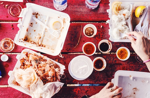 A red outdoor table is covered with various takeout food containers, some of which are empty or almost empty. There are disposable cups with sauces, lemon wedges, rice, and shrimp shells visible. Hands are using plastic cutlery to eat the remaining rice.
