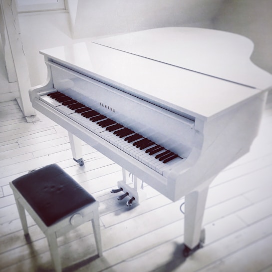A white grand piano with a glossy finish stands on white wooden flooring. The piano's brand is visible on the front. A matching white bench with a dark cushion is positioned next to it. The setting is bright, with light streaming in through a window in the background.