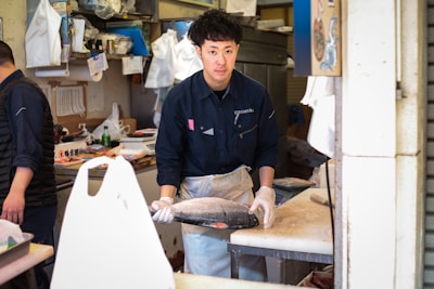 A person is standing in a fish market, wearing a dark blue uniform and gloves, holding a fish on a cutting board. The setting is cluttered with various market supplies and tools such as plastic bags, cutting boards, and boxes.