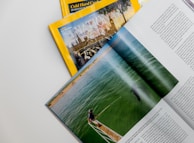 A collection of magazines with a focus on National Geographic, featuring an explorer theme. An open magazine displays an image of a person by the water, possibly involved in a fishing or marine activity, with text on the right side.