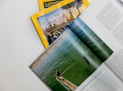 A collection of magazines with a focus on National Geographic, featuring an explorer theme. An open magazine displays an image of a person by the water, possibly involved in a fishing or marine activity, with text on the right side.
