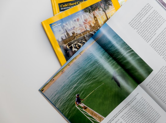 A collection of magazines with a focus on National Geographic, featuring an explorer theme. An open magazine displays an image of a person by the water, possibly involved in a fishing or marine activity, with text on the right side.