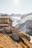 A historic hotel building is perched on the edge of a steep mountain slope. The structure is made of stone and wood, with green shutters and a red sign displaying 'HOTEL' and 'RESTAURANT'. Snow-capped mountains dominate the background under a clear sky. The sunlight casts a warm glow on the landscape, highlighting the contrast between the golden-brown grass and the snow.