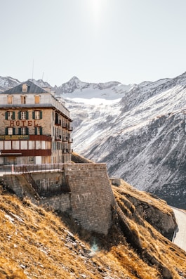 A historic hotel building is perched on the edge of a steep mountain slope. The structure is made of stone and wood, with green shutters and a red sign displaying 'HOTEL' and 'RESTAURANT'. Snow-capped mountains dominate the background under a clear sky. The sunlight casts a warm glow on the landscape, highlighting the contrast between the golden-brown grass and the snow.