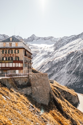 A historic hotel building is perched on the edge of a steep mountain slope. The structure is made of stone and wood, with green shutters and a red sign displaying 'HOTEL' and 'RESTAURANT'. Snow-capped mountains dominate the background under a clear sky. The sunlight casts a warm glow on the landscape, highlighting the contrast between the golden-brown grass and the snow.