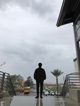 A silhouette of a person stands on a platform overlooking a sports field and multiple vehicles, including a yellow school bus. The sky appears overcast with heavy clouds, and the surroundings include metal railings, a palm tree, and a modern building structure.