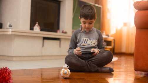 A young boy is seated on a wooden floor inside a cozy room, focused intently on a tablet device. In front of him is a small spherical robot toy, likely BB-8 from Star Wars. The surroundings include a fireplace with decorative items, soft lighting from a window with curtains, and a part of an orange sofa.