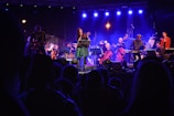 A vibrant live concert scene with musicians performing on stage under blue and purple lighting. The central figure appears to be a singer, surrounded by an ensemble including a guitarist, keyboardist, and several string instrument players. The audience is visible in the foreground, silhouetted against the stage lights.