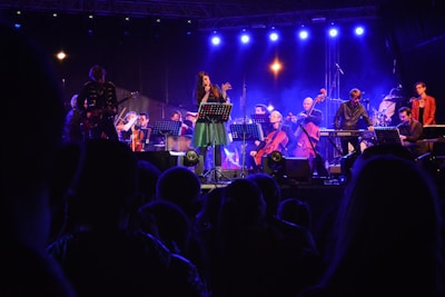 A vibrant live concert scene with musicians performing on stage under blue and purple lighting. The central figure appears to be a singer, surrounded by an ensemble including a guitarist, keyboardist, and several string instrument players. The audience is visible in the foreground, silhouetted against the stage lights.
