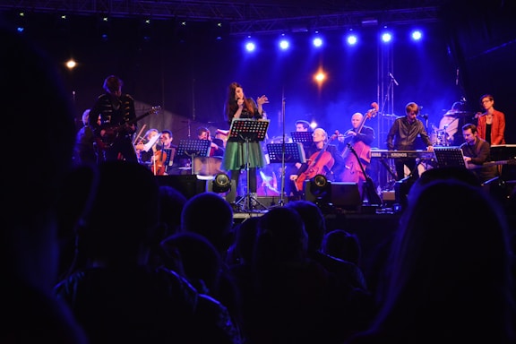 A vibrant live concert scene with musicians performing on stage under blue and purple lighting. The central figure appears to be a singer, surrounded by an ensemble including a guitarist, keyboardist, and several string instrument players. The audience is visible in the foreground, silhouetted against the stage lights.