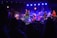 A vibrant live concert scene with musicians performing on stage under blue and purple lighting. The central figure appears to be a singer, surrounded by an ensemble including a guitarist, keyboardist, and several string instrument players. The audience is visible in the foreground, silhouetted against the stage lights.