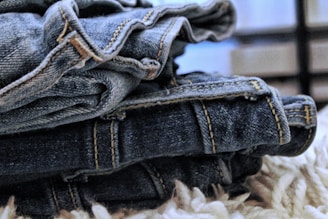 A close-up view of a stack of folded blue denim jeans with visible stitching and texture, resting on a soft, fluffy, light-colored surface.