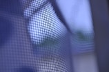 A close-up of a wire mesh fence, in front of a blue-toned blurred background. The metal grid is in clear focus, highlighting its texture and pattern.