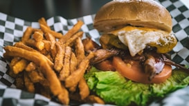 A mouth-watering burger topped with bacon, melted cheese, and a fried egg sits on a bed of fresh lettuce and sliced tomatoes. It is accompanied by a generous serving of crispy sweet potato fries, all placed on a checkered paper background.