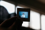 A hand holds up a Kodak transparency slide featuring an image of a man in formal attire, sitting and smiling. The background is softly blurred, highlighting the slide against a bright window in the background.