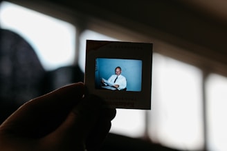 A hand holds up a Kodak transparency slide featuring an image of a man in formal attire, sitting and smiling. The background is softly blurred, highlighting the slide against a bright window in the background.