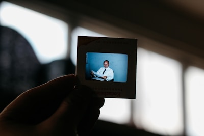 A hand holds up a Kodak transparency slide featuring an image of a man in formal attire, sitting and smiling. The background is softly blurred, highlighting the slide against a bright window in the background.