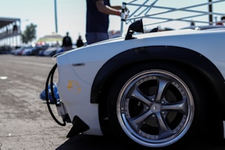A close-up of a car's polished wheel and black tire on pavement, part of a sleek white vehicle body with a decal. The background includes a blurred track setting and silhouettes of people standing near what appears to be a race event.