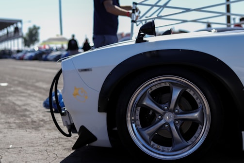 A close-up of a car's polished wheel and black tire on pavement, part of a sleek white vehicle body with a decal. The background includes a blurred track setting and silhouettes of people standing near what appears to be a race event.