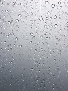 Droplets of water scattered across a smooth glass surface, with varying sizes and clear reflections. The background appears slightly blurred and gray, emphasizing the clarity and texture of the water droplets.